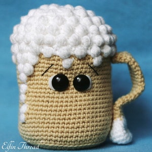 May include: A crocheted beer mug with a white foam top and googly eyes. The mug is beige and has a brown handle.