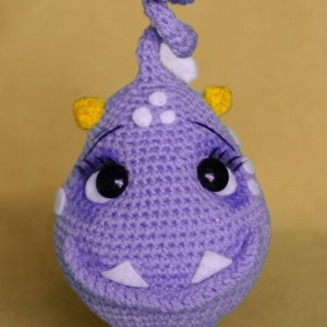 May include: A purple crocheted monster toy with white and yellow accents. The monster has a big smile, big eyes, and a white crocheted flower on top of its head.