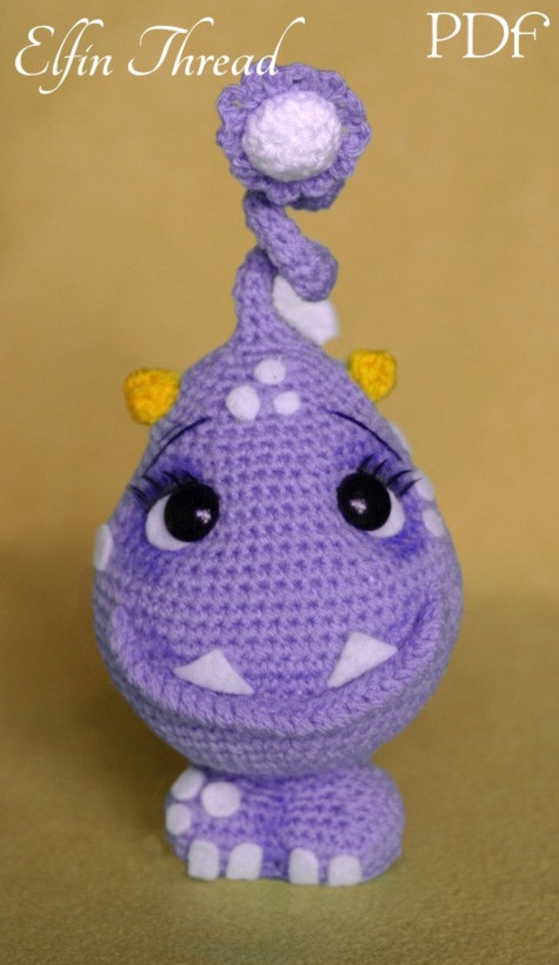May include: A purple crocheted monster toy with white and yellow accents. The monster has a big smile, big eyes, and a white crocheted flower on top of its head.