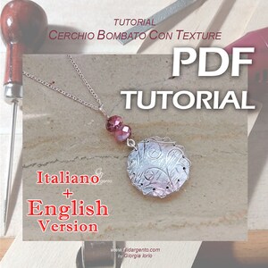Pode incluir: Um colar de prata com um pingente redondo texturizado, duas contas de vidro rosa e uma corrente de prata. O pingente tem um design de espiral e está preso a uma corrente de prata. O colar está sobre uma superfície de madeira com uma lima e uma alça de madeira.