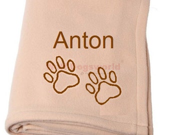Könnte beinhalten: Eine hellrosa Fleece-Decke mit dem Namen Anton, der in Braun gestickt ist. Zwei braune Pfotenabdrücke sind ebenfalls auf die Decke gestickt.