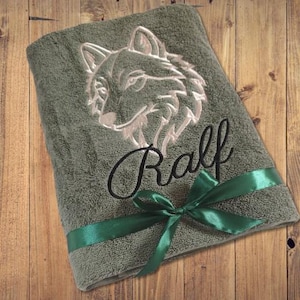 Könnte beinhalten: Ein salbeigrünes Handtuch mit einem gestickten Wolfskopf und dem Namen "Ralf" in Schreibschrift. Ein grünes Satinband ist um das Handtuch gebunden. Das Handtuch liegt gefaltet auf einer Holzoberfläche.