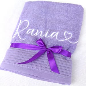 May include: A lavender towel embroidered with the name "Rania" in white script, with a heart symbol. A matching purple satin ribbon is tied around the towel in a bow.