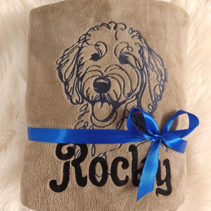Op de afbeelding: Een bruine fleece deken met een blauw lint eromheen gebonden. De deken heeft een geborduurd beeld van een hond met de naam "Rocky" geborduurd onder het beeld.