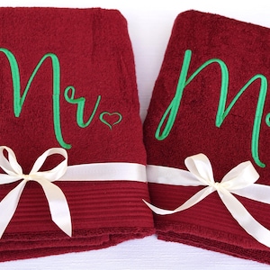 Könnte beinhalten: Zwei weinrote Badetücher, eines mit "Mr" und das andere mit "Mrs" in grüner Schrift bestickt. Jedes Handtuch hat ein weißes Band mit Schleife und ein kleines Herz. Ein Hochzeits- oder Jubiläumsgeschenk.