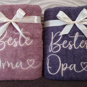 Könnte beinhalten: Zwei flauschige Badetücher, eines in Mauve und eines in Dunkelblau, sind mit weißen Satinbändern gebunden. Das mauvefarbene Handtuch hat den gestickten Text "Beste Oma" und ein Herz. Das dunkelblaue Handtuch hat "Bester Opa" und ein Herz.
