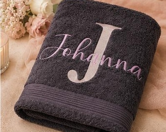 May include: A dark charcoal gray bath towel featuring a textured, raised pattern across its surface. The towel is embroidered with the name Johanna in a light pink script font, with a large, prominent capital letter J in a pale beige tone.