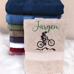 Peut inclure: Serviette de bain beige avec un motif brodé noir et vert d'une personne à vélo de montagne. Le texte "Jürgen" est brodé en vert au-dessus du motif.