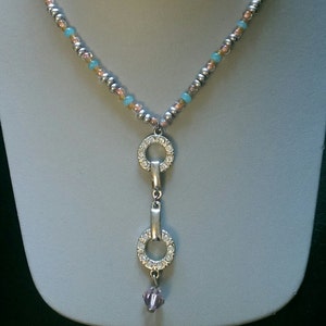 May include: A delicate necklace with a silver pendant featuring two interlocking circles adorned with clear rhinestones. The necklace is made with small, light blue, pink, and silver beads.