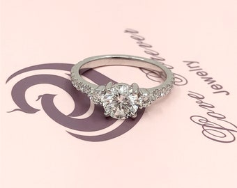 May include: A silver ring with a round, clear center stone and two smaller stones on either side. The band is set with small, clear stones.