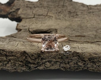 May include: A rose gold engagement ring with a square-shaped, light pink gemstone.