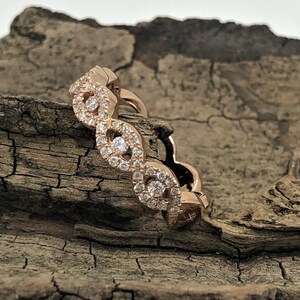 May include: A rose gold band ring with a delicate, intricate design featuring a row of small diamonds set in a wavy pattern.