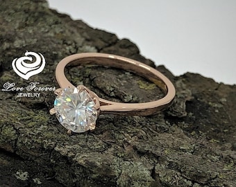May include: A rose gold engagement ring with a large, round, clear center stone. The ring is set on a piece of tree bark.