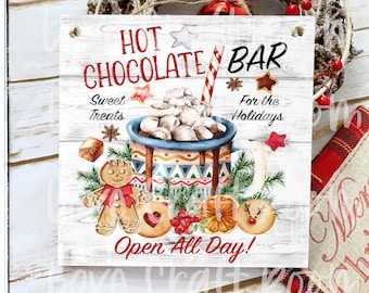 May include: A square wooden sign with the words "Hot Chocolate Bar" in red and black. The sign features a watercolor illustration of a mug of hot chocolate with marshmallows, gingerbread cookies, and other holiday treats. The sign is surrounded by a Christmas wreath.