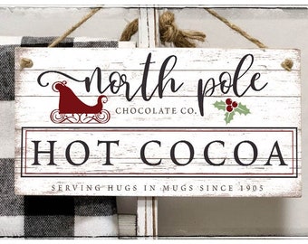 May include: A rectangular wooden sign with the words "North Pole Chocolate Co. HOT COCOA" in black and red lettering. The sign includes a red sleigh graphic and holly leaves. It is suspended by a jute rope.