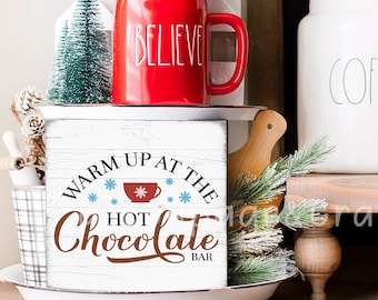 May include: A festive display featuring a red mug with "BELIEVE" in white, a wooden sign that reads "WARM UP AT THE HOT CHOCOLATE BAR", and a small green Christmas tree. The sign has a snowflake and cup design. The items are arranged on a white tray.