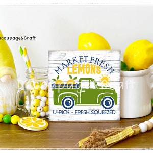 May include: A white wooden sign with the text "Market Fresh Lemons" in blue and green lettering. A green pickup truck with a yellow sun and lemons in the back is pictured on the sign. The text "U-Pick - Fresh Squeezed" is below the truck.