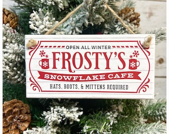 May include: A rectangular wooden sign with the text "Frosty's Snowflake Cafe" in red lettering. The sign also includes the text "Open All Winter" and "Hats, Boots, & Mittens Required". The sign is hanging from a twine string.