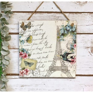 May include: A square wooden sign with a vintage Parisian theme. The sign features a black and white illustration of the Eiffel Tower with a bluebird flying above it. The background is a cream color with a floral pattern and handwritten text. The sign has a rustic, distressed look and is perfect for adding a touch of French charm to any home.