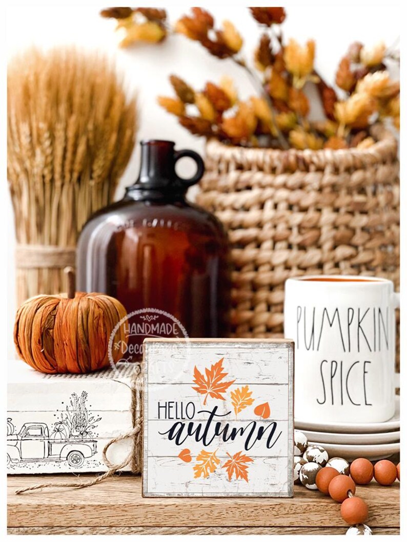 May include: A rustic autumn-themed display featuring a small wooden sign with "Hello Autumn" text and leaf graphics. A pumpkin, a mug with "Pumpkin Spice" text, and other fall decor are also present.