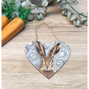 May include: A gray wooden heart-shaped sign with a brown rabbit illustration. The sign has a white twine hanger and features the text "Happy Easter" and "Happy Eggs".