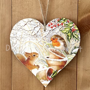 May include: A wooden heart-shaped ornament featuring a winter scene with a robin perched on a pot and a squirrel peeking out from behind a bush. The background is a snowy forest with holly berries.