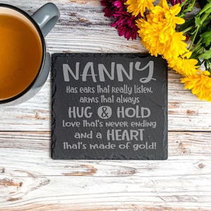 May include: A black slate coaster with the word "NANNY" engraved at the top, accompanied by a loving message. A cup of tea and yellow and purple flowers are arranged around the coaster.
