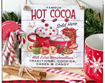 May include: A wooden sign with the text "FAMOUS HOT COCOA SOLD HERE" features a red mug with whipped cream and candies. The sign is surrounded by candy canes, cinnamon sticks, and a red mug with a snowflake design. The sign also says "With Extra Marshmallows, Traditional Cookies, Cakes & Candy."