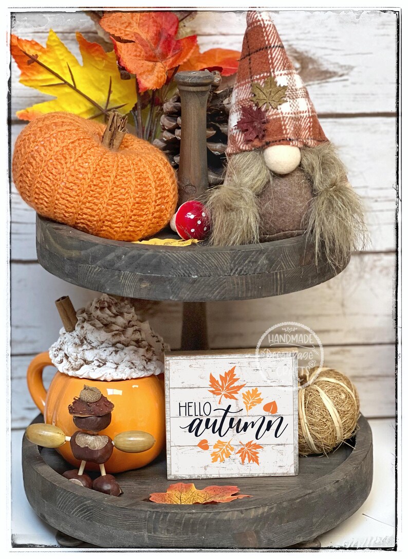 May include: A two-tiered wooden tray with fall-themed decorations. Includes an orange pumpkin, a gnome with a plaid hat, a mug with whipped cream, and a sign that says "Hello Autumn".