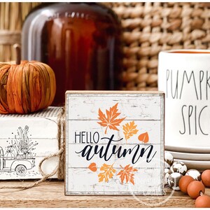 May include: A rustic autumn-themed display featuring a decorative wooden sign with "Hello Autumn" text and orange leaf accents. A white mug with "Pumpkin Spice" text, a decorative pumpkin, and other fall decor are also present.