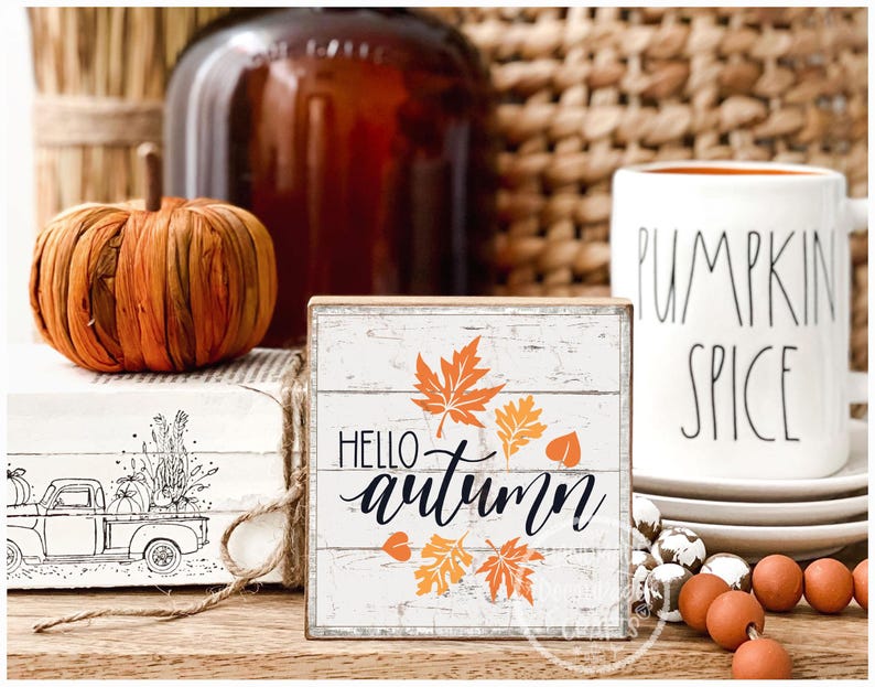 May include: A rustic autumn-themed display featuring a decorative wooden sign with "Hello Autumn" text and orange leaf accents. A white mug with "Pumpkin Spice" text, a decorative pumpkin, and other fall decor are also present.