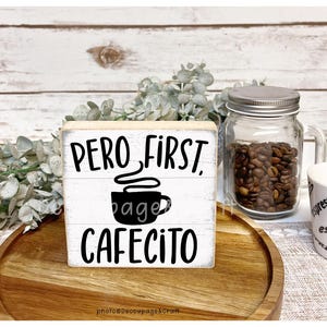 Peut inclure: Un panneau en bois blanc avec le texte "Pero, First, Cafecito" en noir. Un dessin de tasse à café noire est au centre. Un bocal en verre rempli de grains de café et une tasse à expresso blanche sont également visibles.