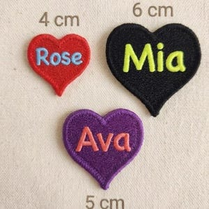 May include: Three embroidered heart-shaped patches. The red heart, 4 cm wide, has "Rose" in blue. The black heart, 6 cm wide, has "Mia" in yellow. The purple heart, 5 cm wide, has "Ava" in pink. Each patch has a stitched border.