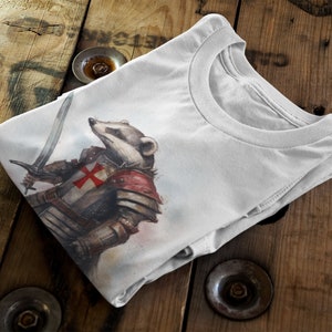 Pode incluir: Camiseta branca com uma ilustra&ccedil;&atilde;o em aquarela de um texugo em armadura medieval, segurando uma espada. A armadura inclui uma cruz vermelha no peito. A camiseta est&aacute; dobrada sobre uma superf&iacute;cie de madeira.