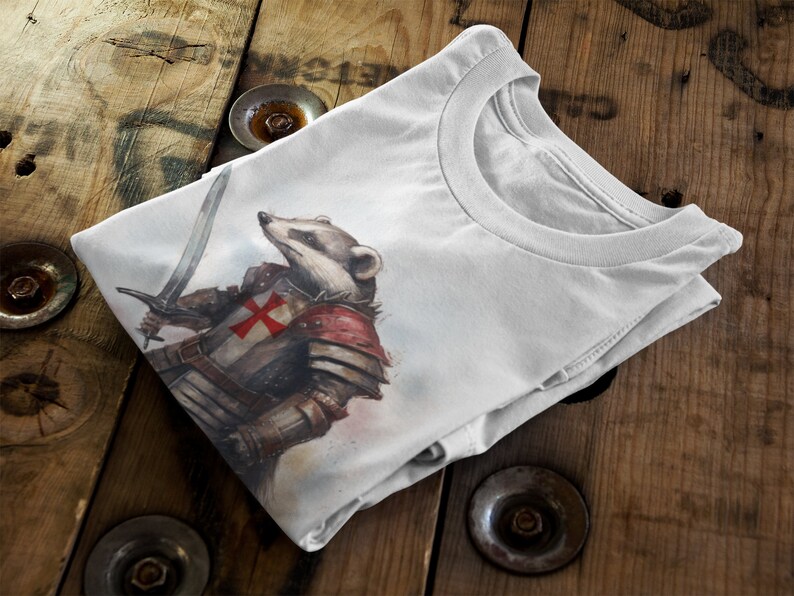 Pode incluir: Camiseta branca com uma ilustra&ccedil;&atilde;o em aquarela de um texugo em armadura medieval, segurando uma espada. A armadura inclui uma cruz vermelha no peito. A camiseta est&aacute; dobrada sobre uma superf&iacute;cie de madeira.