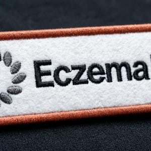 Op de afbeelding: Een rechthoekige geborduurde patch met een witte viltachtergrond en een koperkleurige gestikte rand. Links een cirkelvormig logo gemaakt van kleine grijze geborduurde ovalen. Rechts de zwarte geborduurde tekst EczemaUK.