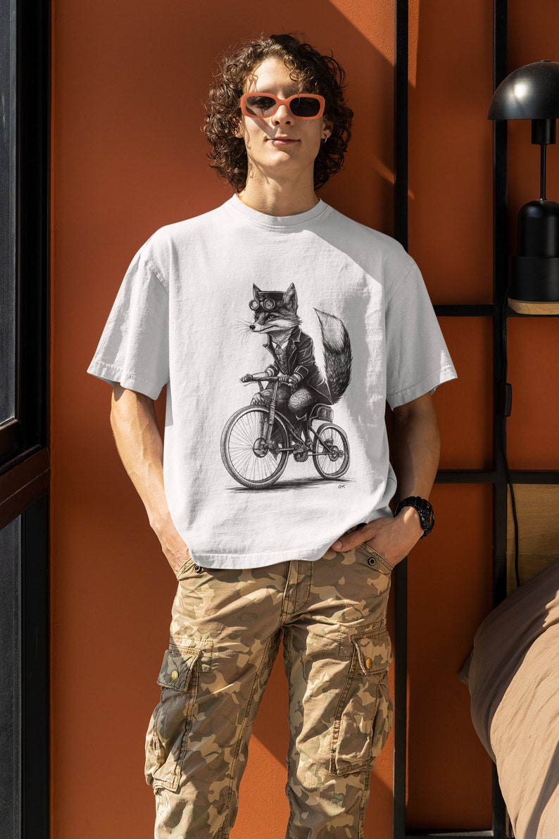 以下が含まれることがあります： ゴーグルを着用し、自転車に乗っているキツネの白黒イラストが描かれた白いTシャツ。キツネはジャケットを着ており、尻尾があります。