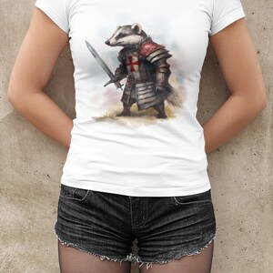 Pode incluir: Camiseta branca com uma ilustra&ccedil;&atilde;o em aquarela de um texugo com armadura de cavaleiro, segurando uma espada. O texugo usa um capacete e uma cruz vermelha no peito. A camiseta &eacute; combinada com shorts jeans pretos.