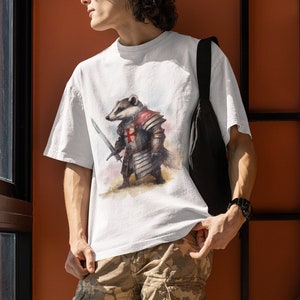 Pode incluir: Camiseta branca com uma ilustra&ccedil;&atilde;o em aquarela de um texugo em armadura medieval, segurando uma espada. A armadura inclui uma cruz vermelha no peito. A pessoa est&aacute; usando cal&ccedil;as cargo camufladas e uma bolsa de ombro preta.