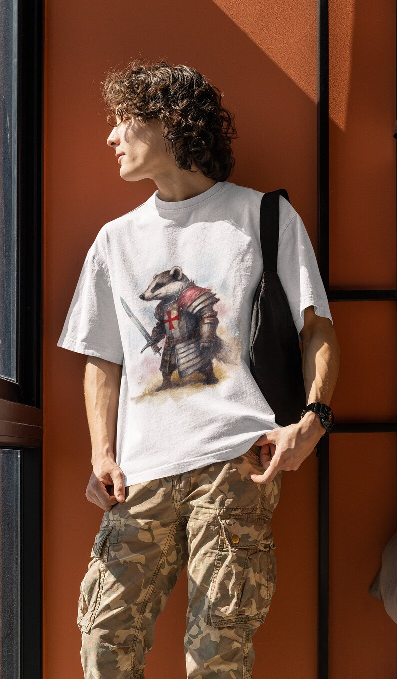 Pode incluir: Camiseta branca com uma ilustra&ccedil;&atilde;o em aquarela de um texugo em armadura medieval, segurando uma espada. A armadura inclui uma cruz vermelha no peito. A pessoa est&aacute; usando cal&ccedil;as cargo camufladas e uma bolsa de ombro preta.