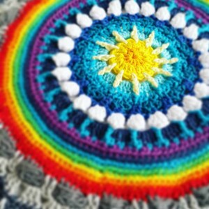 Puede incluir: Un mandala de crochet colorido con un borde de arcoíris y un sol amarillo en el centro.