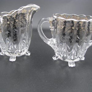 May include: A vintage glass creamer and sugar bowl set with silver accents. The set features a floral design and has a footed base.