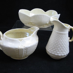 May include: A set of three vintage Irish Belleek porcelain pieces in a cream and yellow color. The set includes a creamer, a sugar bowl, and a pitcher. The pieces are decorated with a shell pattern.