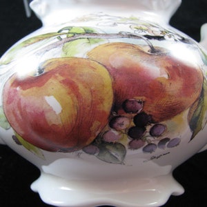 May include: White ceramic teapot with a hand-painted design of red apples, green leaves, and purple grapes. The teapot has a rounded body and a spout on the left side. The handle is on the right side and is attached to the lid.