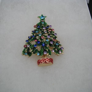 May include: A Christmas tree brooch with green enamel branches and colorful jewel accents. The tree is set in a gold-tone pot with a red and gold design. A star tops the tree.