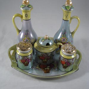 May include: A vintage porcelain condiment set with a blue and green tray. The set includes two cruets, a sugar bowl, and two salt and pepper shakers. The cruets are labeled "O" and "V" and feature floral designs. The sugar bowl has a lid and features a floral design with red apples in baskets. The salt and pepper shakers also feature floral designs with red apples in baskets.
