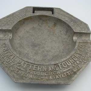 May include: A gray, octagonal metal ashtray featuring embossed text that reads: WOOD AND METAL PATTERNS, ALUMINUM AND BRASS CASTINGS, CENTRAL PATTERN AND FOUNDRY CO., QUALITY PRODUCTION SERVICE, 3737 SO. SACRAMENTO AVE. - LA FAYETTE 4232 - CHICAGO.