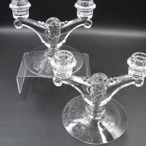May include: Two clear glass double-arm candle holders featuring a central pedestal base, curved arms extending to two candle cups, and decorative beaded detailing along the top edges of the arms, crafted from transparent pressed glass.