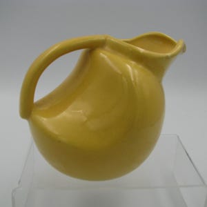 May include: A yellow ceramic pitcher with a curved handle and a rounded body. The pitcher has a spout for pouring and is displayed on a clear surface. The pitcher is a solid, bright yellow color.