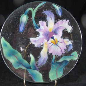 May include: A glass plate with a painted design of a purple orchid with green leaves. The plate has a textured surface and a dark background.